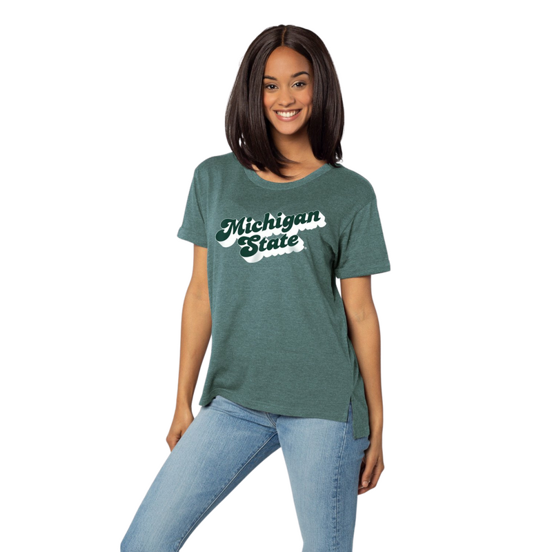 Woman wearing a retro style t-shirt in a faded green with short sleeves and a wide crewneck. Back of the t-shirt is slightly longer than the front hem. Printed on the center chest is script font reading Michigan State in dark green with a white shadow 