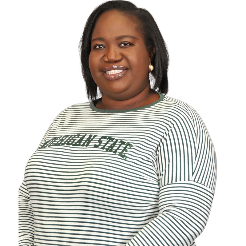 Woman standing at a three-quarter angle, wearing a long-sleeved crewneck t-shirt. The shirt is white with thin green horizontal stripes. The sleeve hem is halfway down the upper arm. Green printing in the center chest reads Michigan State