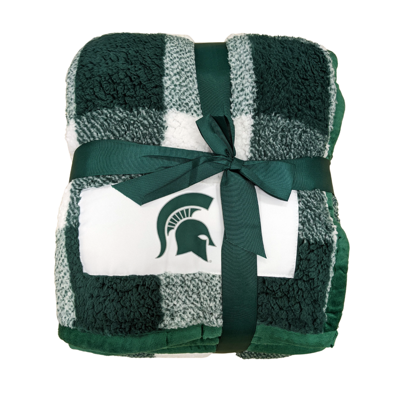 Green and white buffalo plaid fuzzy blanket wrapped into a neat square and tied with a dark green ribbon. On one corner is an embroidered green Spartan helmet, and the blanket's edging a piped dark green fabric.