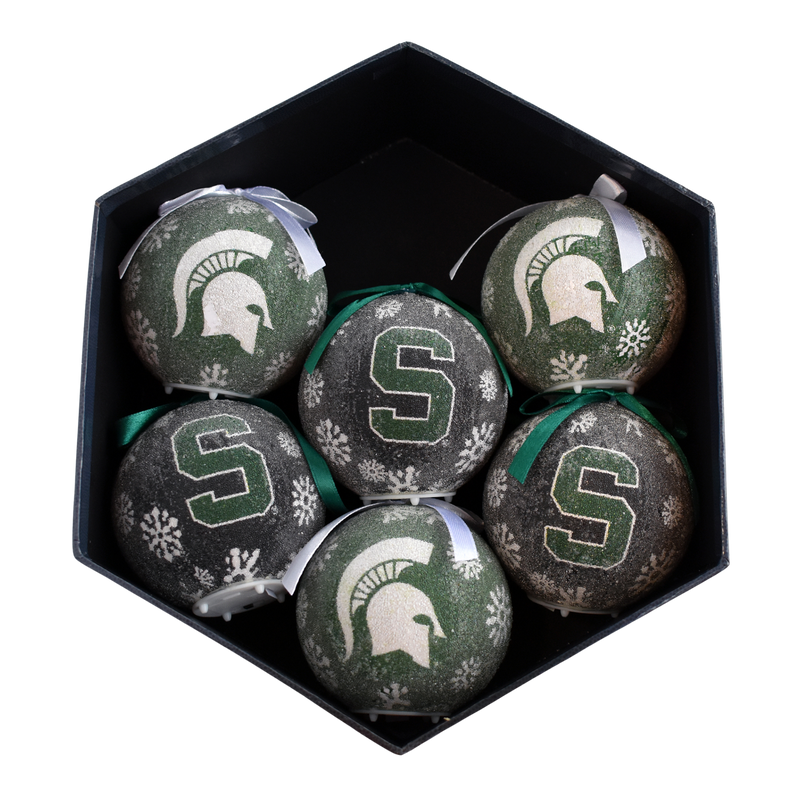 Black hexagonal storage box holding six round, textured ornaments with ribbons. Three are black with white snowflakes, a green block S, and green ribbons. The other three are forest green with white snowflakes, a white Spartan helmet, and a white ribbon.