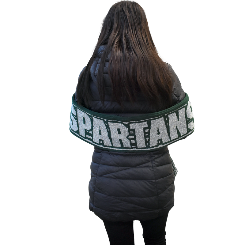 Back view of a woman in a black puffy coat with the dark green and silver striped scarf held by her elbows across her back, displaying the large silvery block text that reads Spartans.