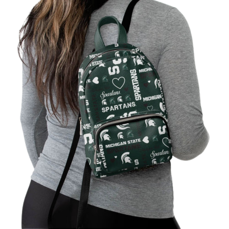 Woman with the mini backpack slung over one shoulder. The backpack is forest green with various Michigan State logos and heart icons in white and black.