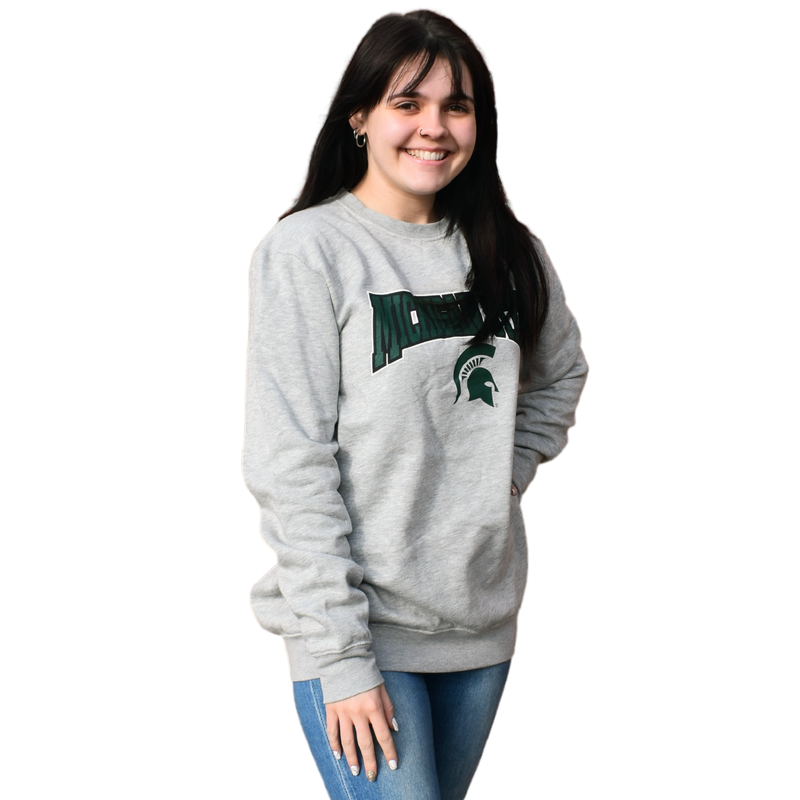 Woman wearing a light heather gray crewneck sweatshirt, with Michigan State printed in a graphic arch over a Spartan helmet on the center chest.