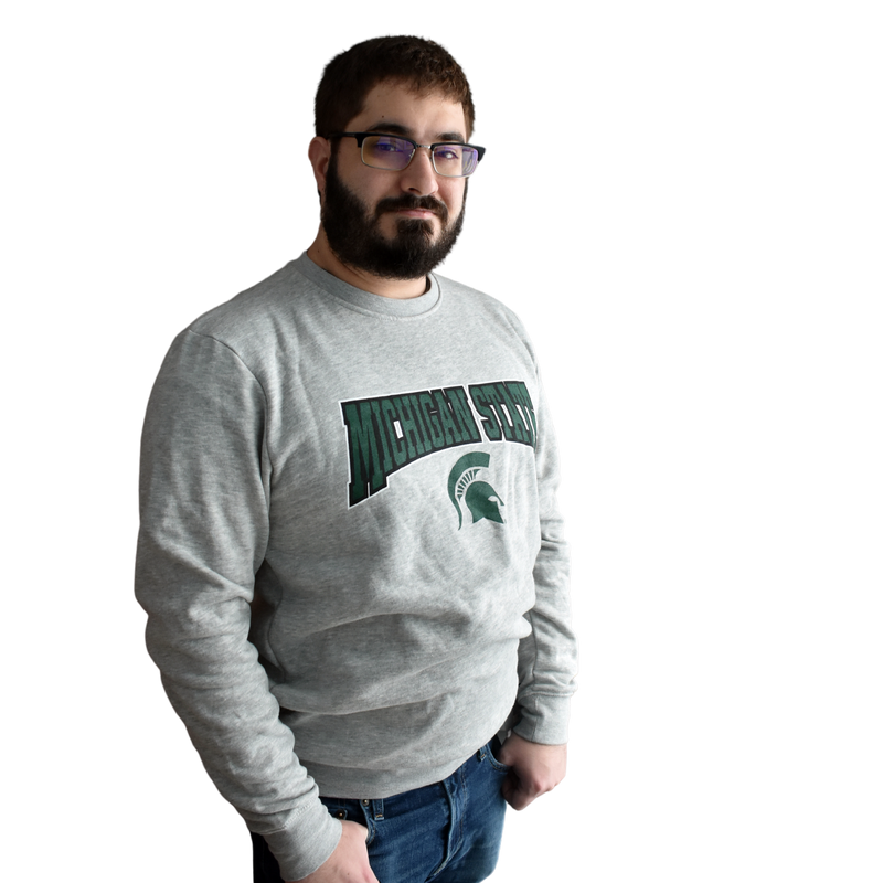 Man wearing a light heather gray crewneck sweatshirt, with Michigan State printed in a graphic arch over a Spartan helmet on the center chest.