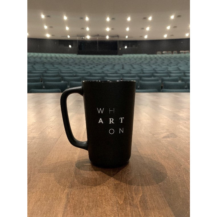 A black coffee mug on the stage of the Wharton Center. The mug has the word Wharton spelled in a staggered formation. 