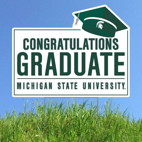 White lawn sign reading "Congratulations, Graduate: Michigan State University" in forest green all caps. Extending off of the top right corner is a forest green graduation cap with a silver tassel and white Spartan helmet.